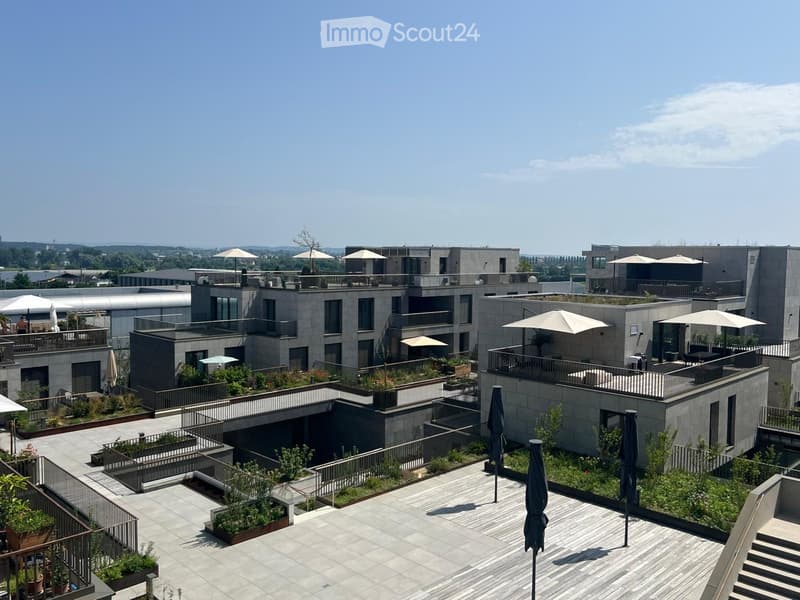 Modern rooftop garden with seating areas, umbrellas, and greenery, surrounded by contemporary buildings.