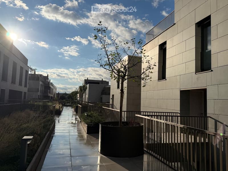 Modern residential complex courtyard with a central tree in a planter, metal railings, and sleek buildings in the background.