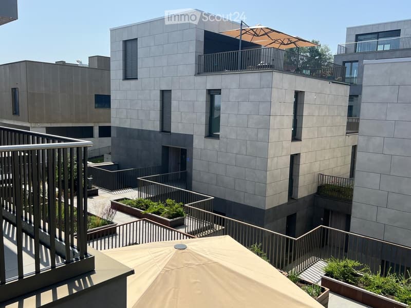 Modern multi-story building with grey stone facade, multiple balconies, and rooftop terrace with umbrella and small garden.