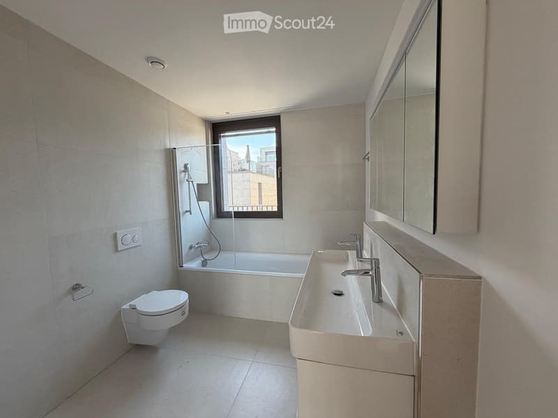 Modern bathroom with a toilet, sink, bathtub, and large mirror. Features a window with a view of a building and balcony.
