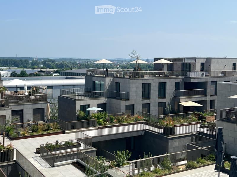 Modern multi-story residential building with balconies, outdoor seating areas, and greenery on the rooftop.