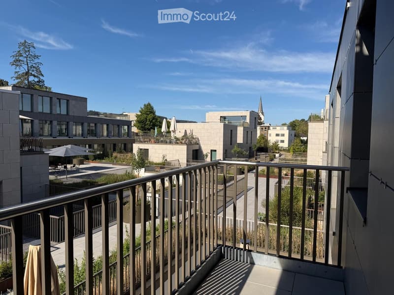 Balcony overlooking a modern residential complex with contemporary buildings, greenery, and outdoor seating areas.