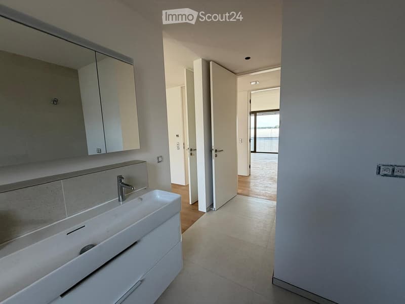 Modern bathroom with a white sink, chrome faucet, mirrored cabinet, and tiled walls. The hallway leads to another room with wooden flooring.