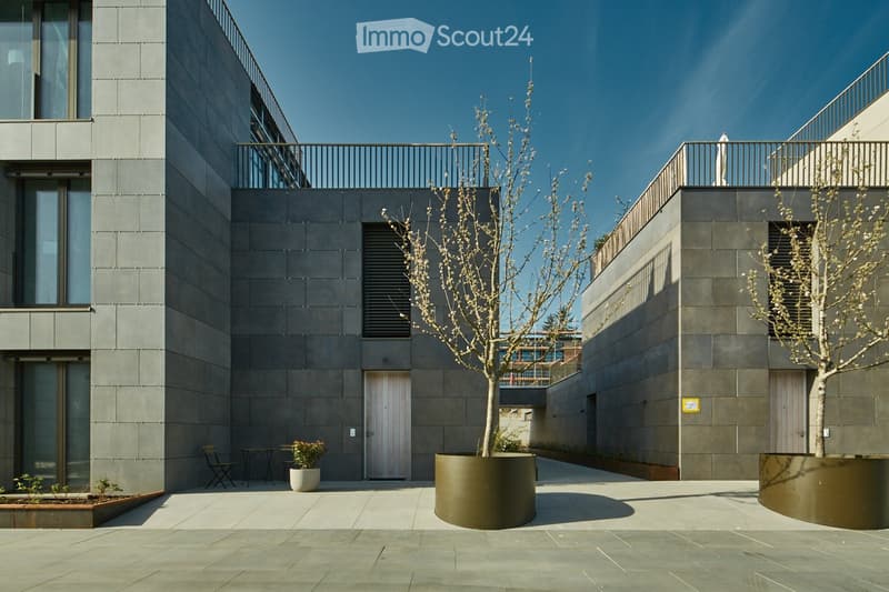 Modern courtyard with grey stone walls, a small tree in a large planter, a small table and chair, and a potted plant.