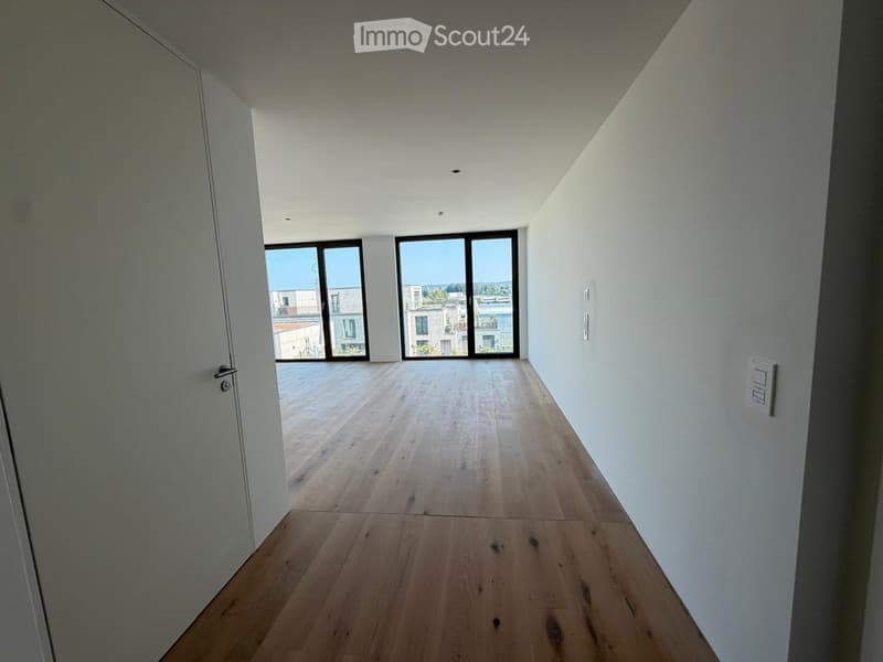 Empty hallway with wooden flooring, white walls, and large windows at the end leading to an outdoor area.