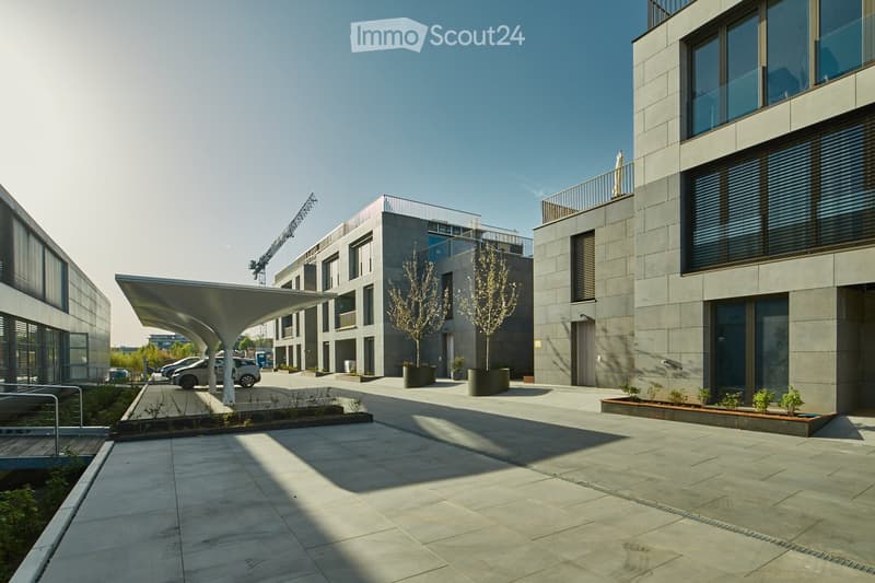 Modern multi-story building with grey stone facade, large windows, and balconies. The courtyard features a canopy structure, small trees, and a paved walkway.