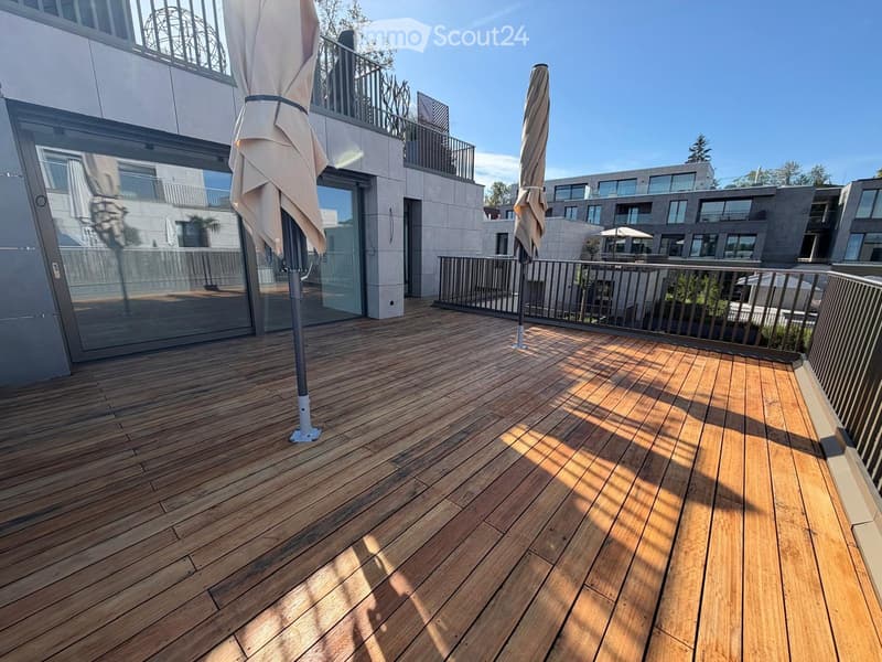 Wooden deck with glass railing, two folded umbrellas, and a modern building in the background.