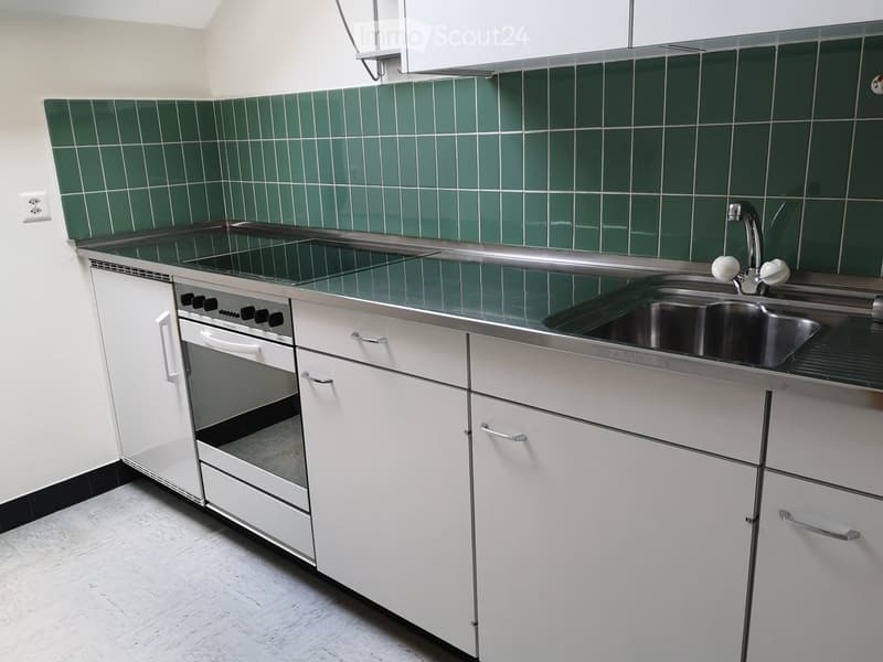 Modern kitchen with white cabinets, stainless steel sink, and built-in oven. Green tiled backsplash and light-colored countertops.