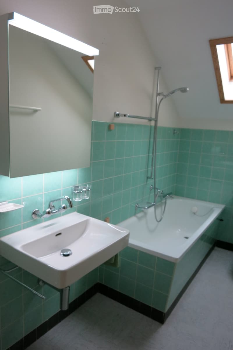 Modern bathroom with a white sink, bathtub, and green tiled walls. Includes a faucet, showerhead, and small cups on the wall.