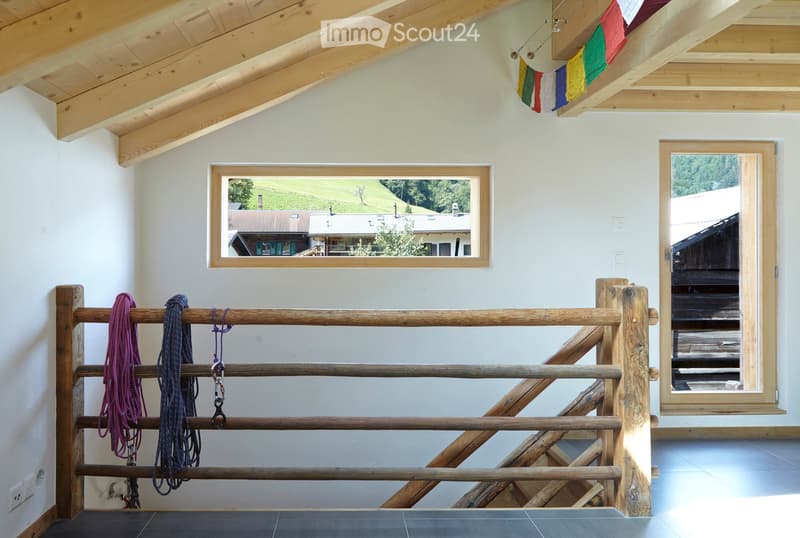 Bright hallway with wooden beams and exposed ceiling, featuring a large window showing a scenic view, a wooden railing with ropes, and colorful flags hanging from the ceiling.