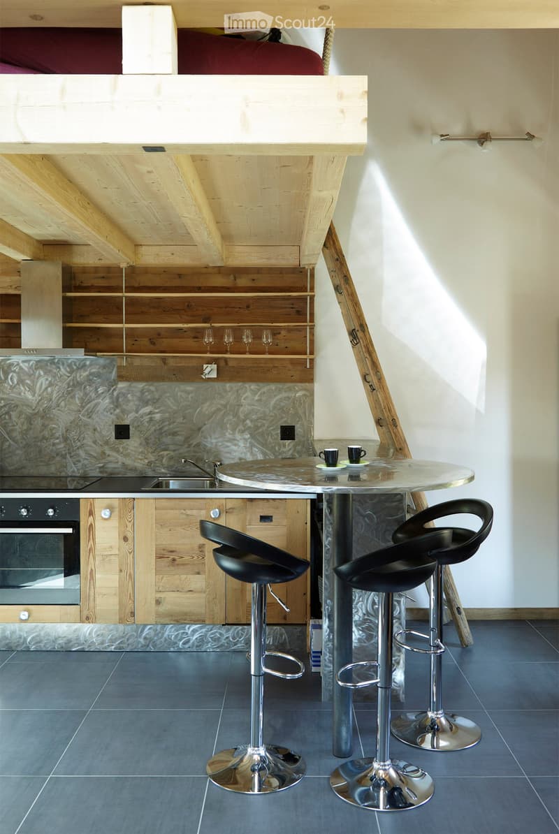 Modern kitchen with wooden ceiling beams, bar table, bar stools, oven, sink, and wine glasses on shelves.
