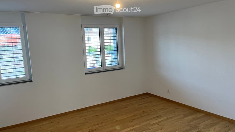 Empty room with light-colored walls, wooden flooring, and two windows with blinds.