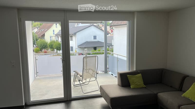 Living room with a grey sofa, green cushions, and a sliding glass door leading to a balcony with a chair.