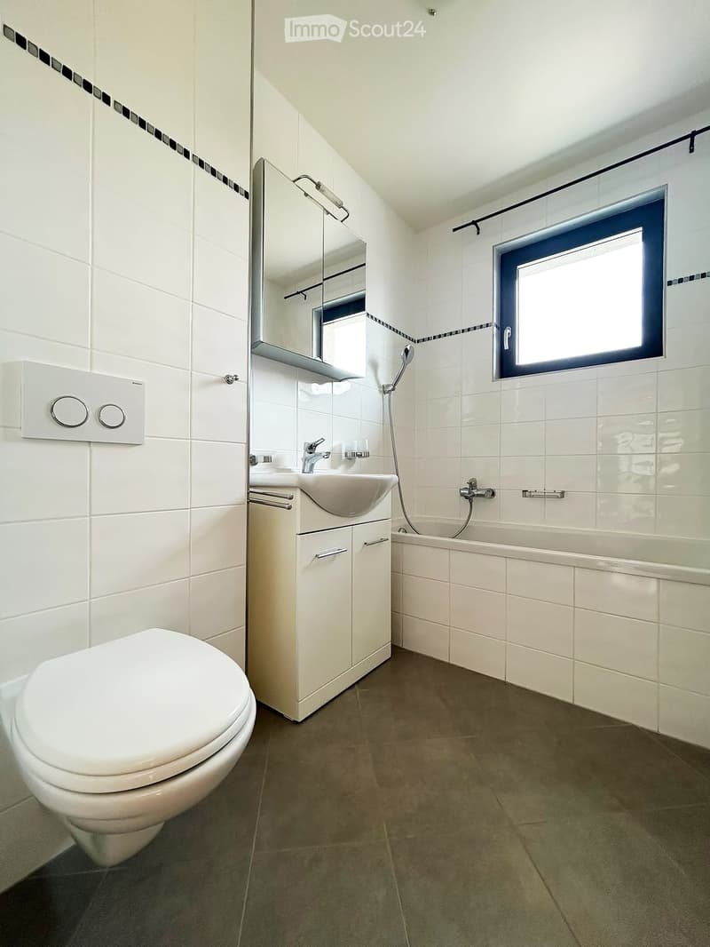 Modern bathroom with white tiles, a toilet, a sink with a cabinet, a bathtub, and a mirror above the sink. The room features a window with a blue frame and a dark grey tiled floor.