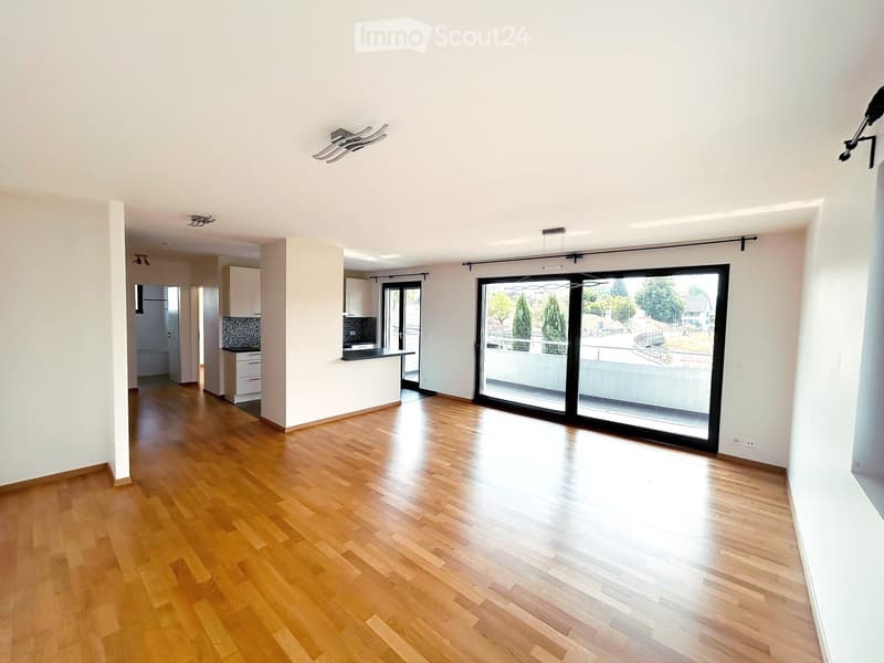 Bright, empty room with wooden flooring, white walls, and large sliding glass doors leading to a balcony. Minimalist design with recessed lighting and a small kitchen area visible in the background.