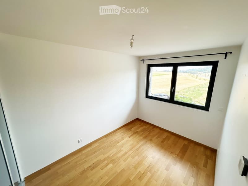 Empty room with light-colored walls, wooden flooring, and a large window offering a view of an outdoor landscape.