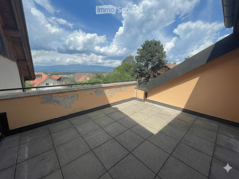 Balcony with tiled floor, low wall, and railing, overlooking a landscape with trees and distant mountains.