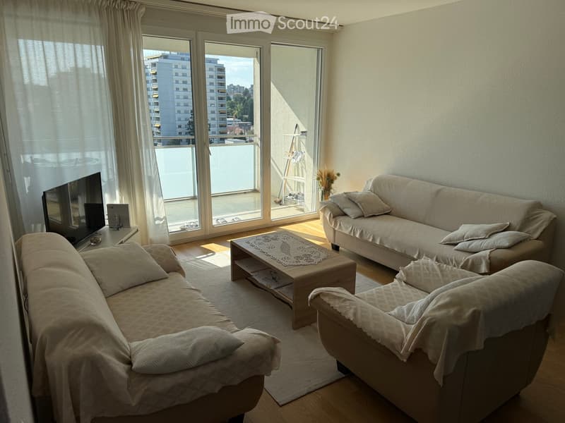 Bright living room with large windows and balcony access, featuring a sofa, armchair, coffee table, and television.