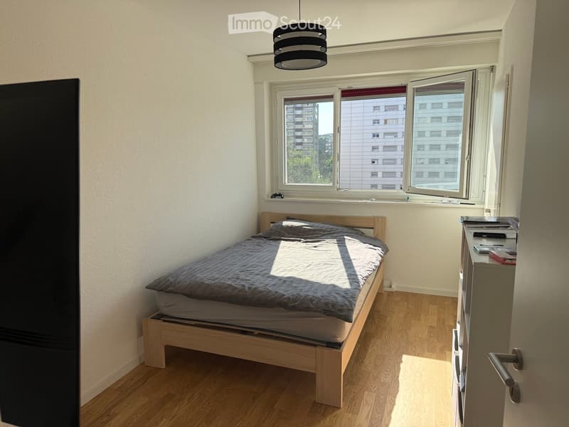 A small, well-lit bedroom with a wooden bed, nightstand, and a hanging lamp. The room features large windows with a view of a building outside.
