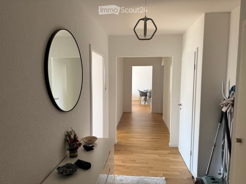 A modern hallway with light wooden flooring, white walls, and a hanging geometric light fixture. A round mirror is mounted on the left wall, and a small console table holds decorative items. The hallway leads to an open area with a dining setup visible in