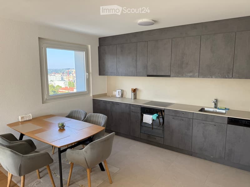 Modern kitchen with grey cabinets, a wooden dining table, four chairs, and a view of the city through a large window.