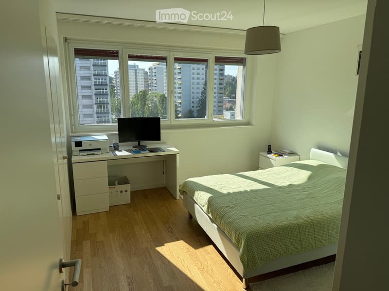 Bright bedroom with wooden flooring, large windows overlooking a cityscape, a neatly made bed, a desk with a computer and printer, and a nightstand with a lamp.