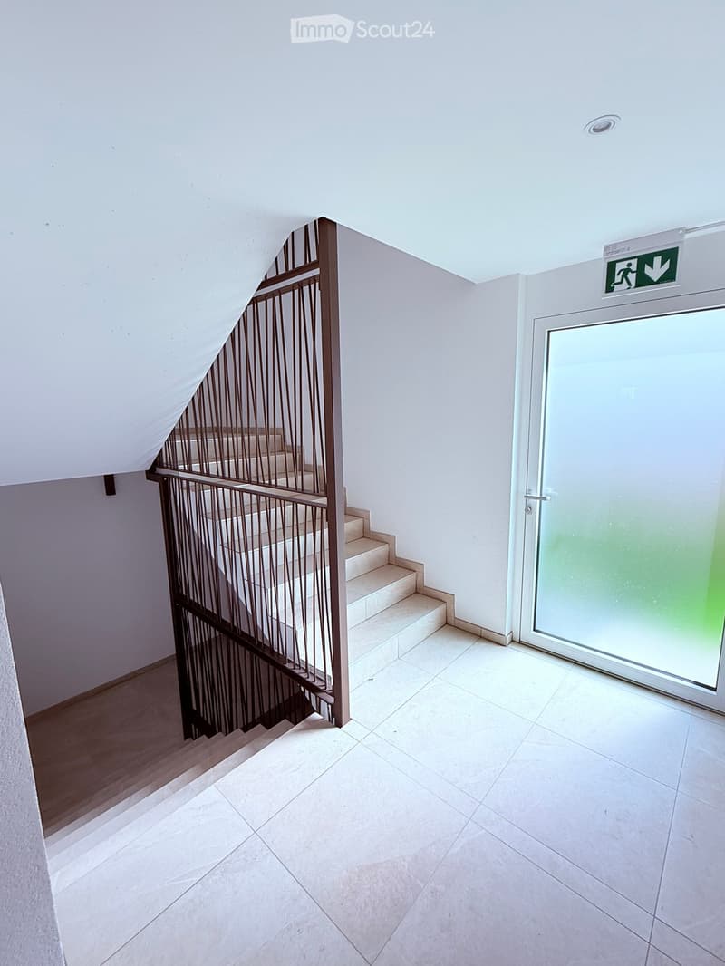 Modern hallway with a staircase featuring a metal railing and beige tiled steps, leading to a glass door with an emergency exit sign above it.