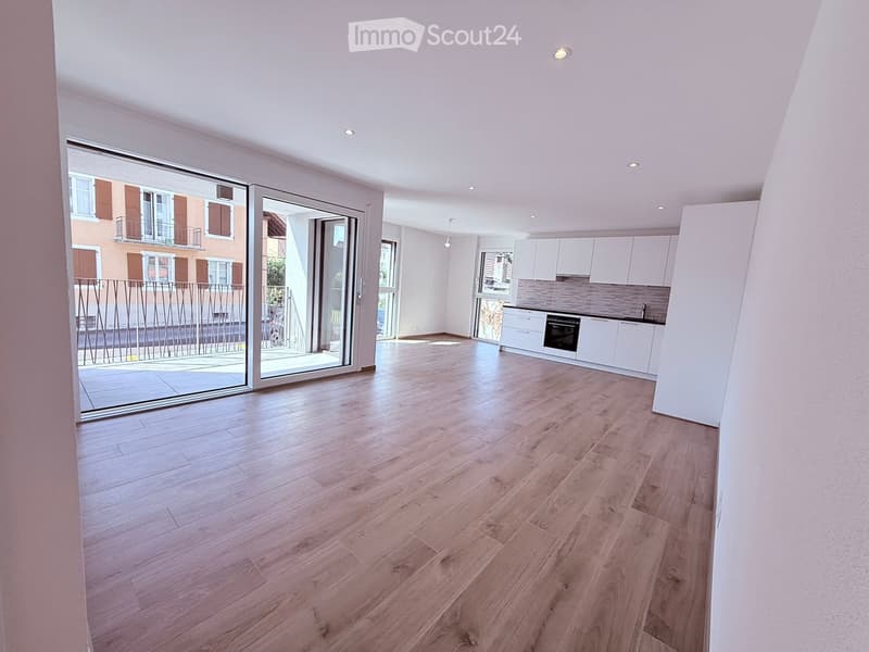 Bright, unfurnished living space with wooden flooring, white walls, recessed lighting, and a large sliding glass door leading to a balcony. Adjacent kitchen area with white cabinets and stainless steel appliances is visible.