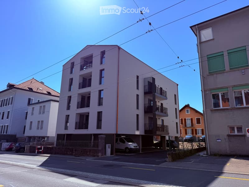 Modern multi-story residential building with balconies, located on a street with other buildings and parked cars.