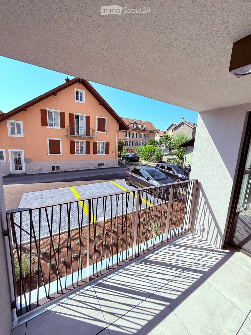 Balcony with metal railing, tiled floor, and small garden area with plants. View of street, parked cars, and neighboring houses.
