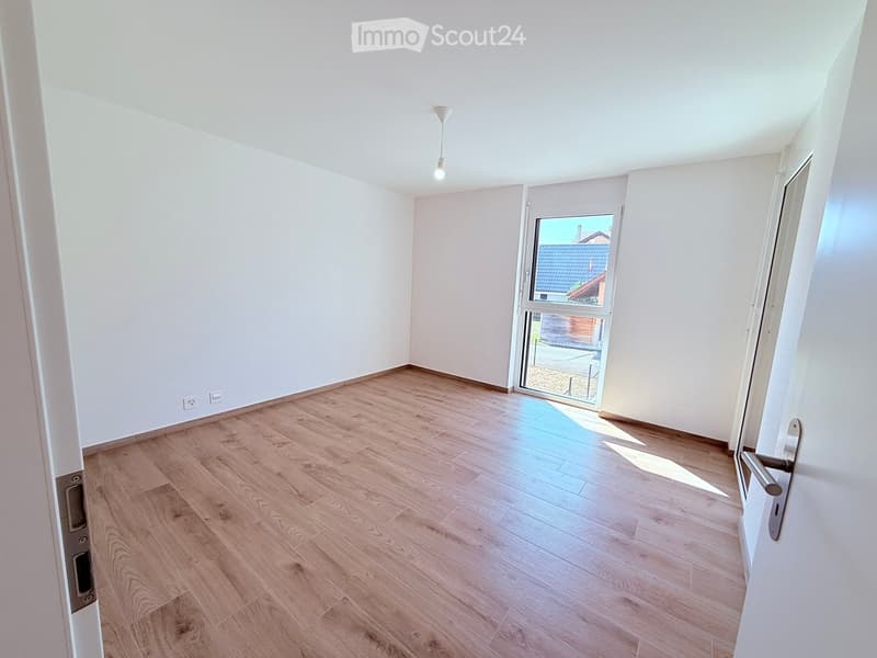 Empty room with light wooden flooring, white walls, and a large window with a view of a neighboring roof.