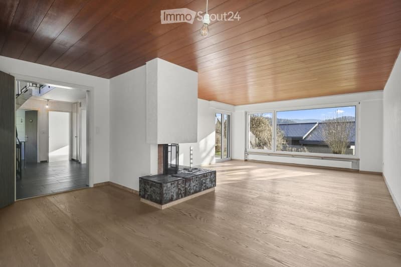 Bright, open living space with wooden floors, white walls, and large windows offering a view of the outdoors. Features a central fireplace and a pendant light fixture.