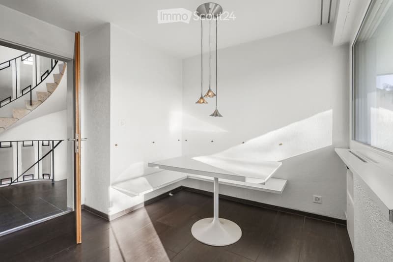 Modern, minimalist room with white walls, dark wooden floor, white table and chair, pendant lights, and a view of a staircase through an open doorway.