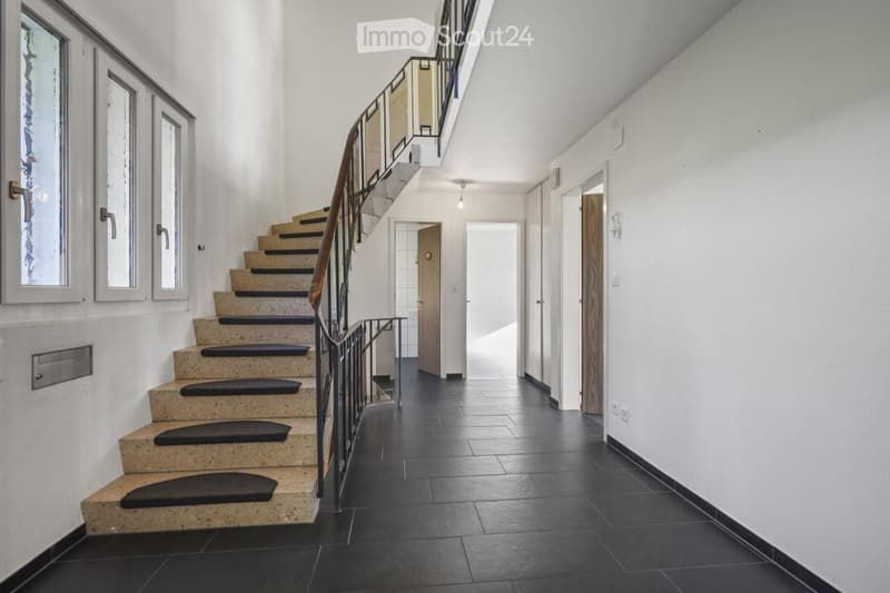 Modern hallway with black tiled floor, white walls, staircase with beige steps and black railings, several doors and windows.