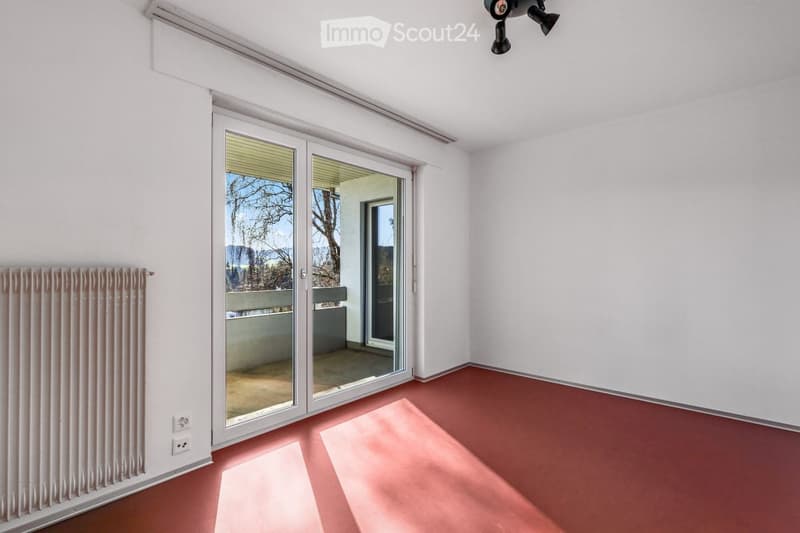 Empty room with white walls, red floor, large sliding glass door leading to a balcony, radiator, and ceiling lights.