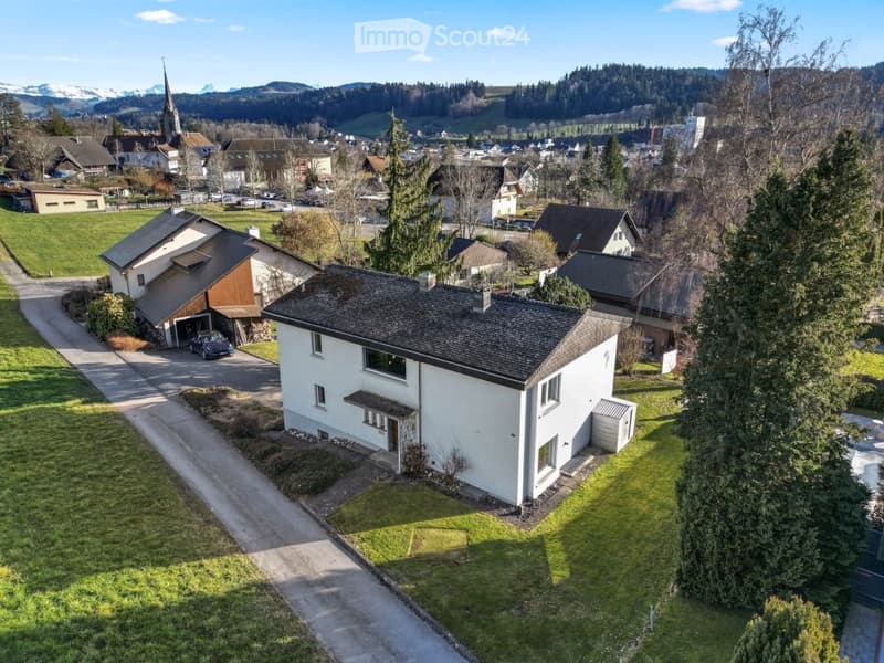 Aerial view of a two-story white house with a dark roof, surrounded by a well-maintained lawn and trees. A driveway leads to a garage with a parked car. The house is situated in a rural area with neighboring houses and a church visible in the background.