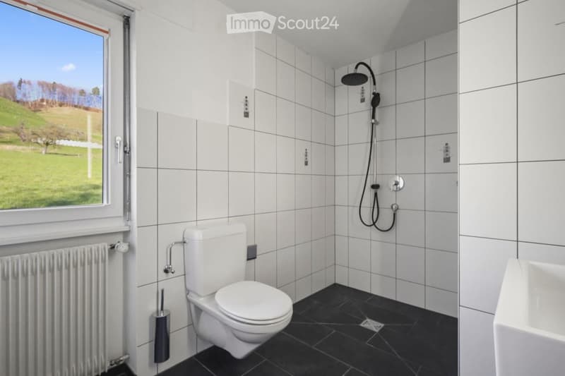 Modern bathroom with white tiled walls, black tiled floor, toilet, shower, and a view of a green landscape through a large window.