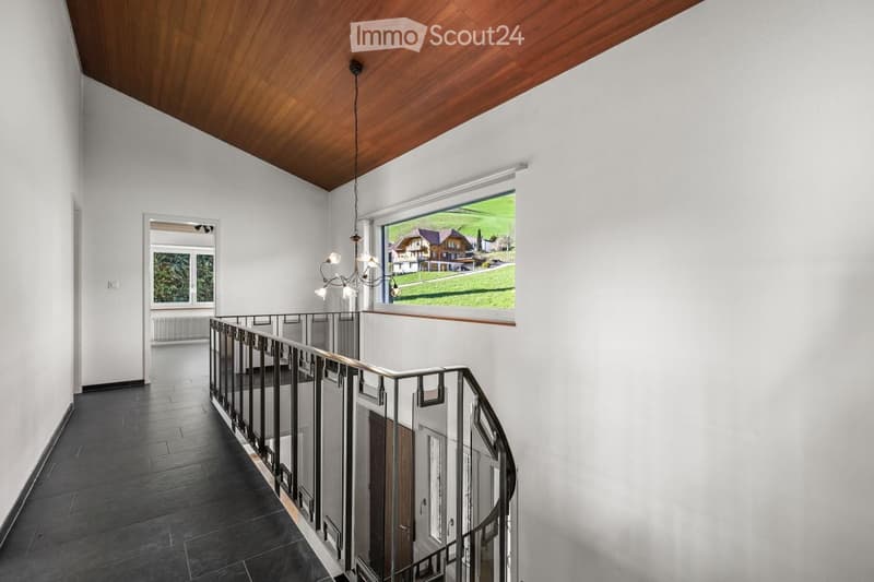Spacious hallway with a wooden ceiling, dark tiled floor, and a modern metal railing leading to an upper level. Large windows offer a view of a green landscape and houses.
