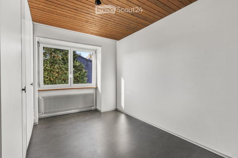 Empty room with white walls, grey concrete floor, wooden ceiling, large window with view of greenery, radiator below window, and a door on the left side.