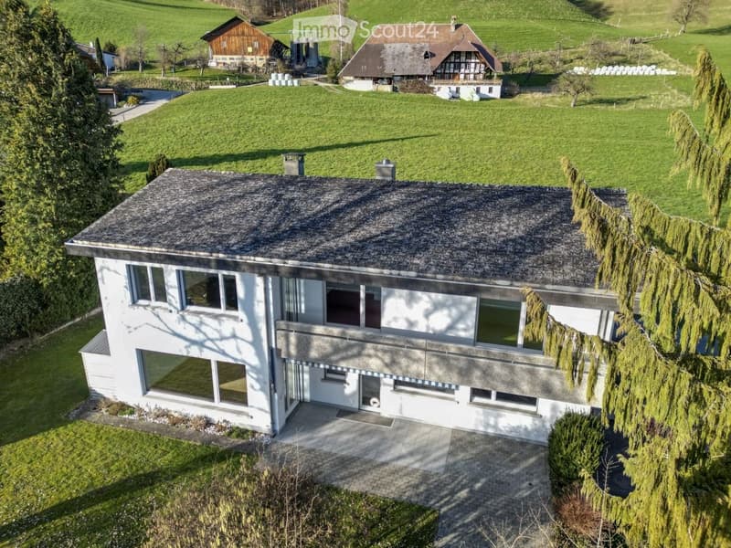 A modern two-story house with a flat roof and large windows, surrounded by green fields and trees. The house features a covered entrance and is situated in a rural landscape with additional buildings visible in the background.