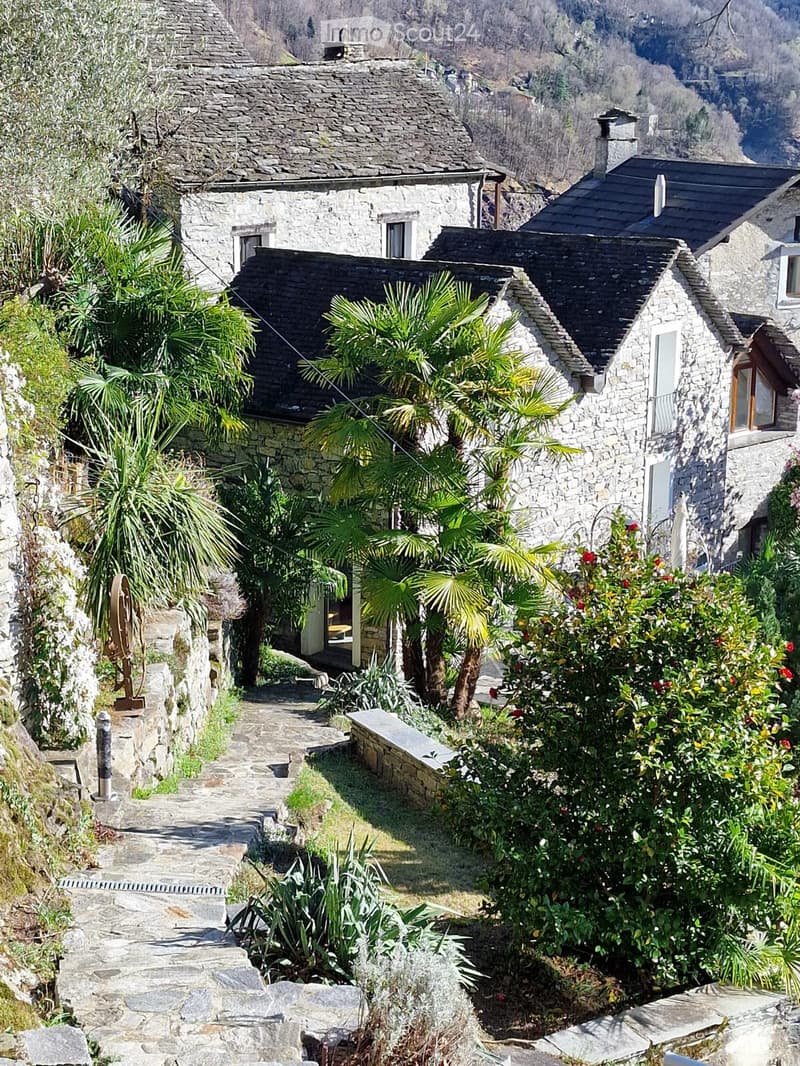 A stone pathway leads up to a rustic stone house with a tiled roof, surrounded by lush greenery and palm trees.