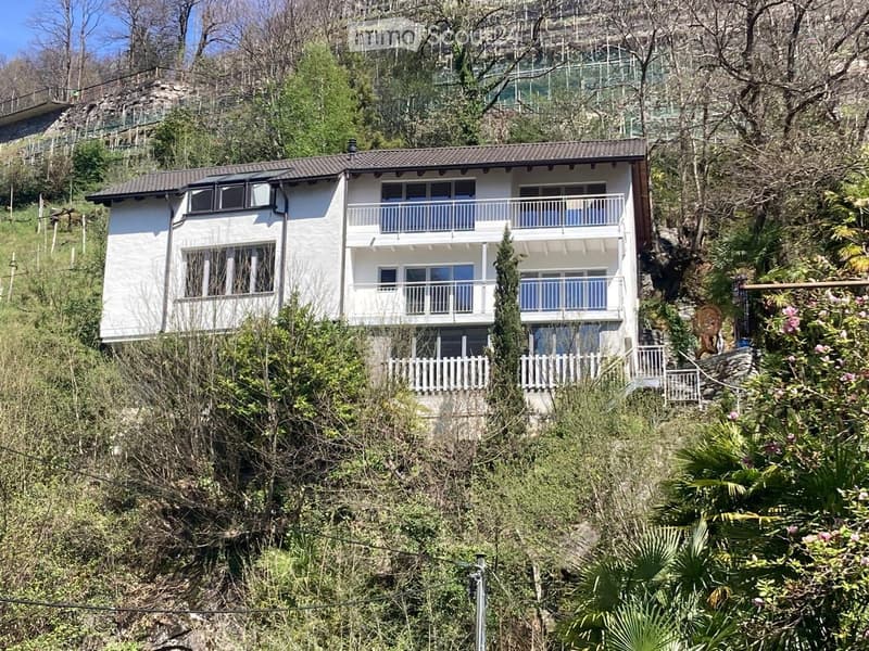 A two-story white house with multiple balconies, situated on a hillside surrounded by greenery and trees.