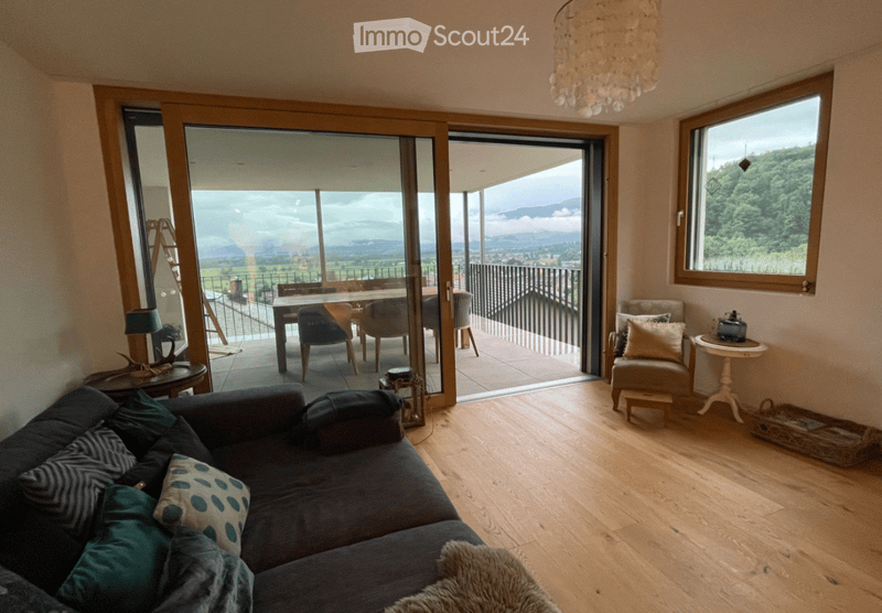 Modern living room with large sliding glass doors opening to a balcony, wooden flooring, a dark grey sectional sofa, a small coffee table, a side table with a lamp, and a dining area with a table and chairs.
