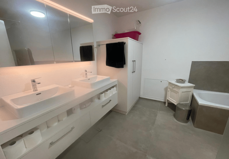 Modern bathroom with double sinks, large mirror, built-in cabinet, towel rack, bathtub, and small stool.