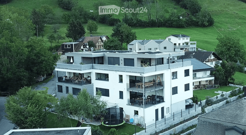 Modern multi-story building with balconies, surrounded by greenery and neighboring houses.