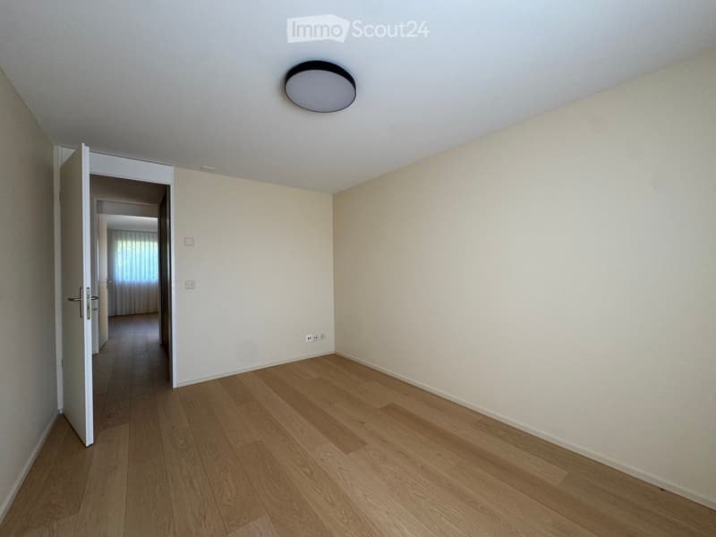 Empty room with light-colored walls, wooden flooring, and a ceiling light fixture. An open doorway leads to another room with visible furniture.