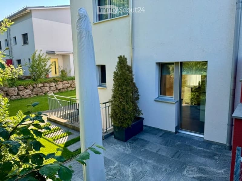 A modern outdoor patio area with a potted tree, stone steps leading to a grassy garden, and a white house in the background.