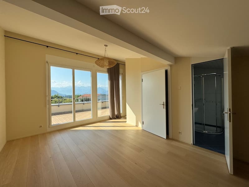 Bright, empty room with wooden flooring, large windows, and a view of the mountains. Features a hanging light fixture and an open doorway to a shower area.