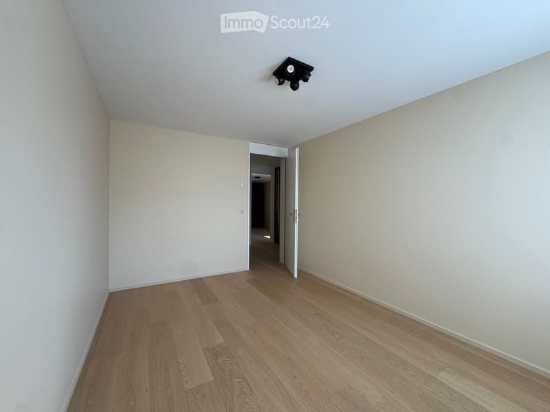 Empty room with light-colored walls and wooden flooring, featuring a ceiling light fixture and an open doorway leading to another area.