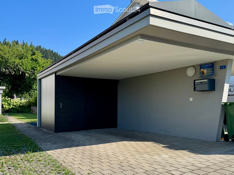 Modern building with a covered entrance, garage door, and mailbox. The entrance features a sleek, flat roof and a clean, contemporary design.